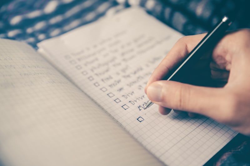 A hand in the foreground uses a pen to write a checklist in an out-of-focus notebook.