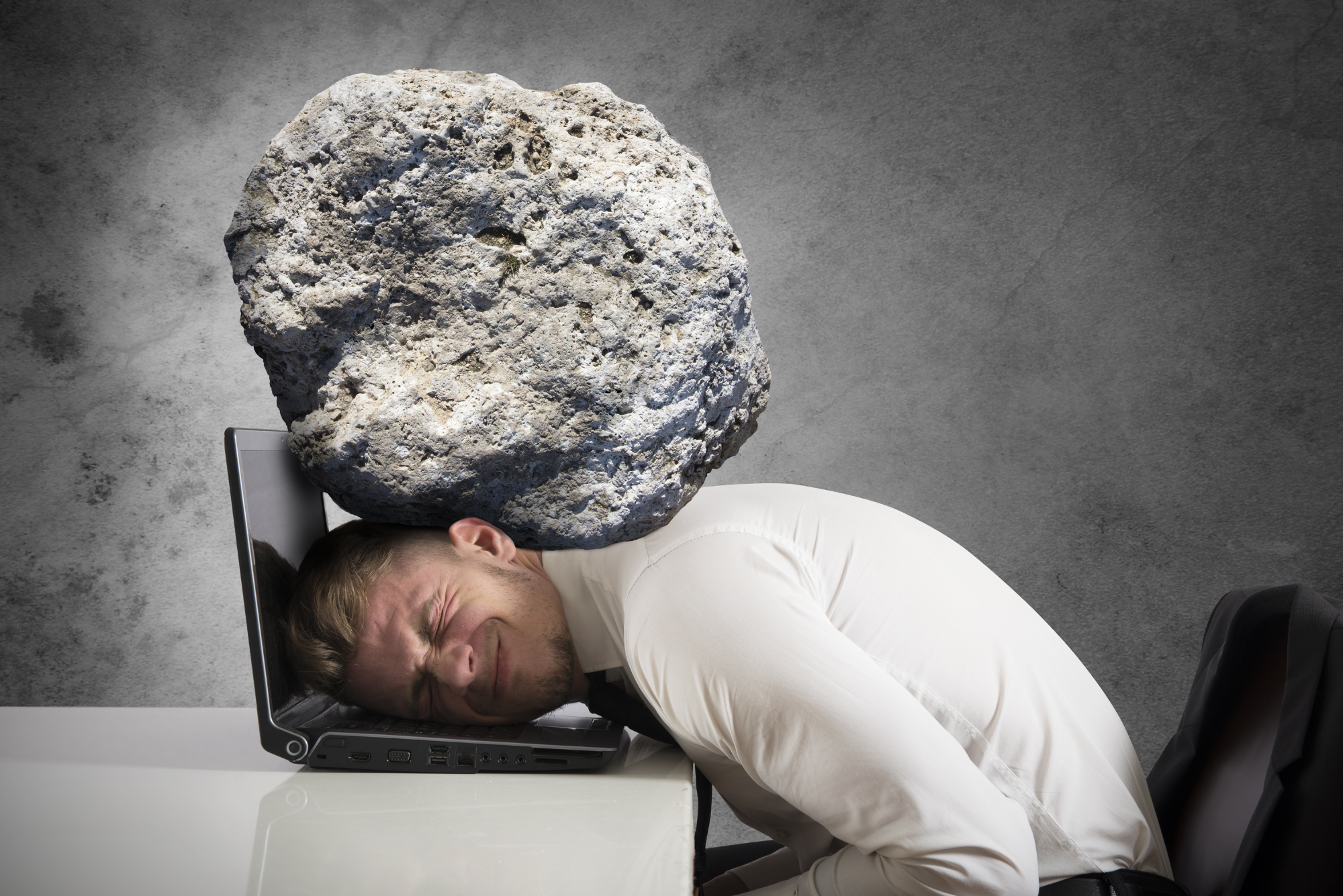 A man lays his head on his laptop with a boulder crushing his head, representing his high stress and desire for a simpler life.