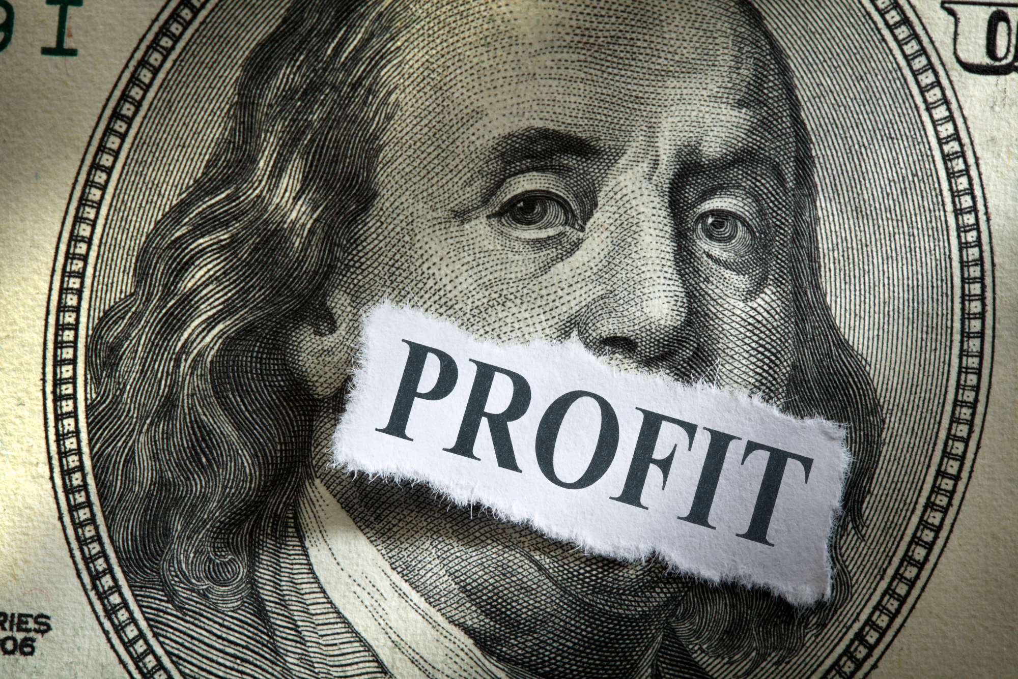 A scrap of paper with the word “profit” on it covers Benjamin Franklin’s mouth on a close-up $100 bill.