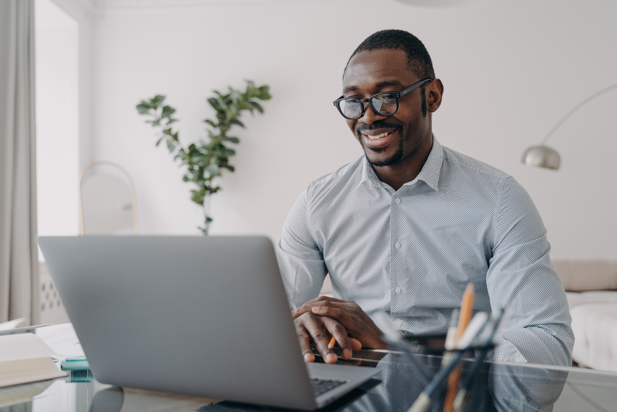 A smiling man wearing glasses and a light shirt while working on a laptop at a desk.