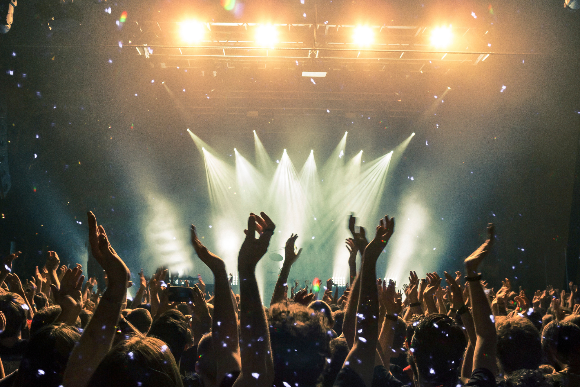 Huge crowd with raised hands at concert with confetti and bright lights.