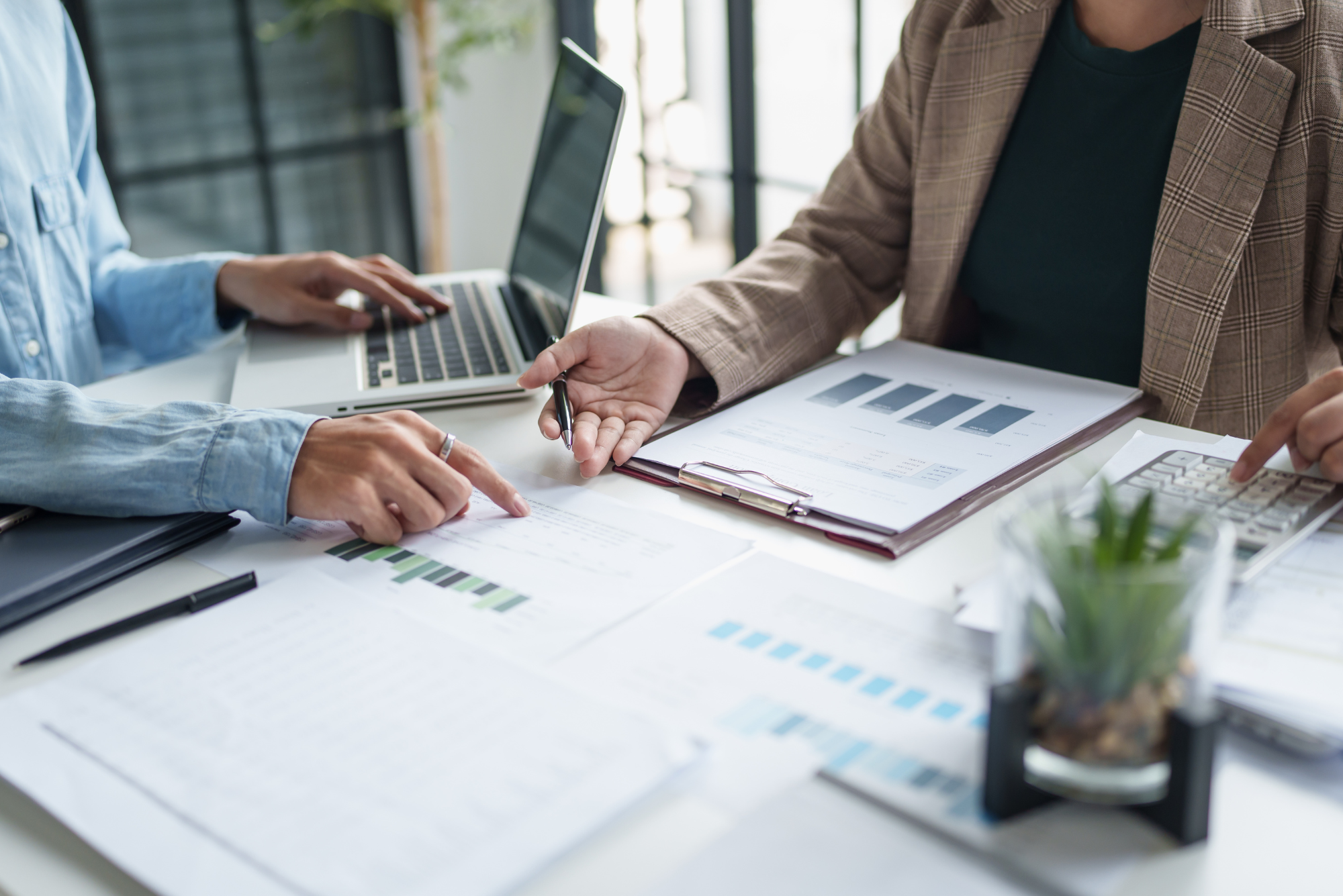 Two business people analyze financial data on papers and laptop during meeting in modern office with large windows.
