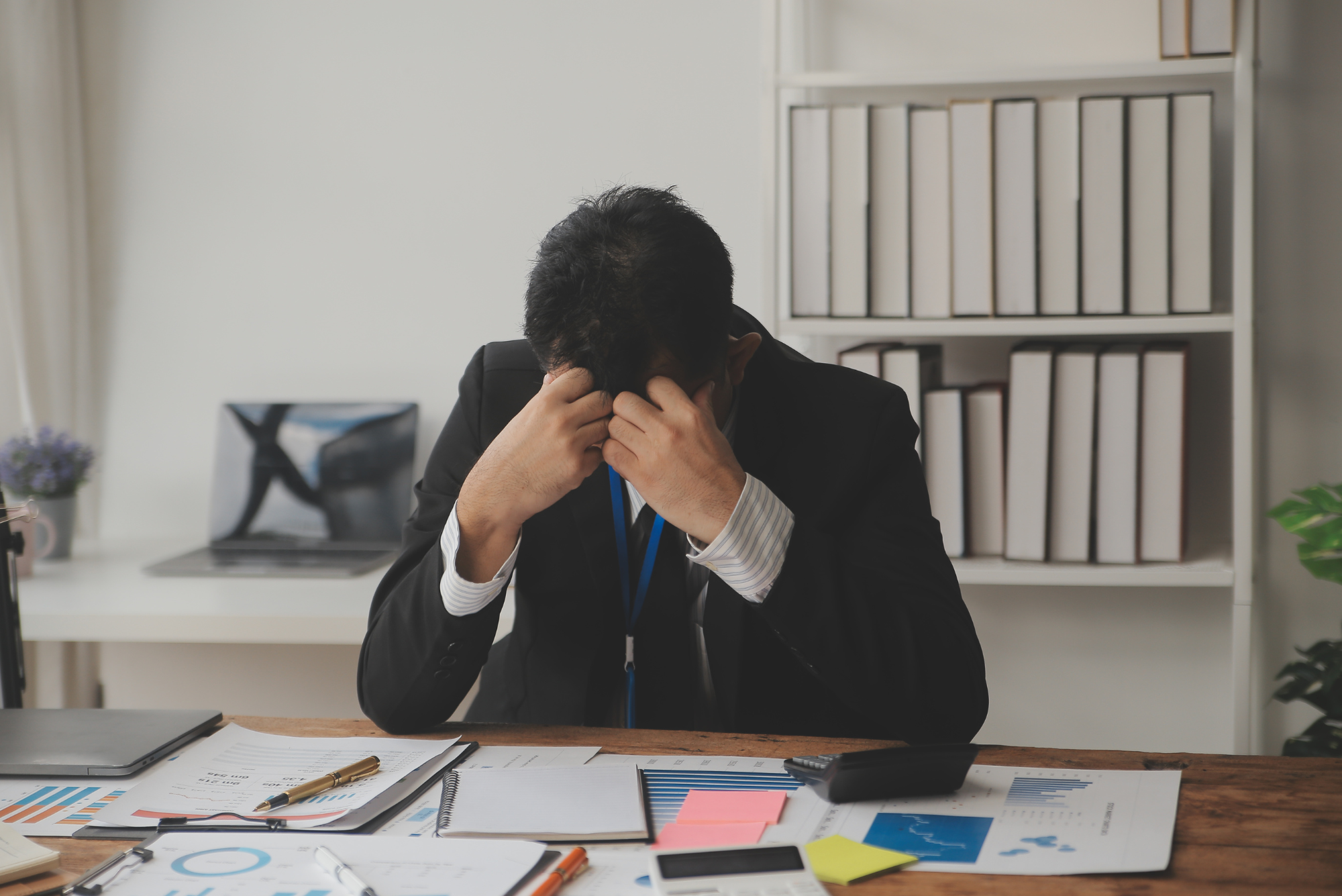 Frustrated businessman with head in hands at messy office desk covered in papers.