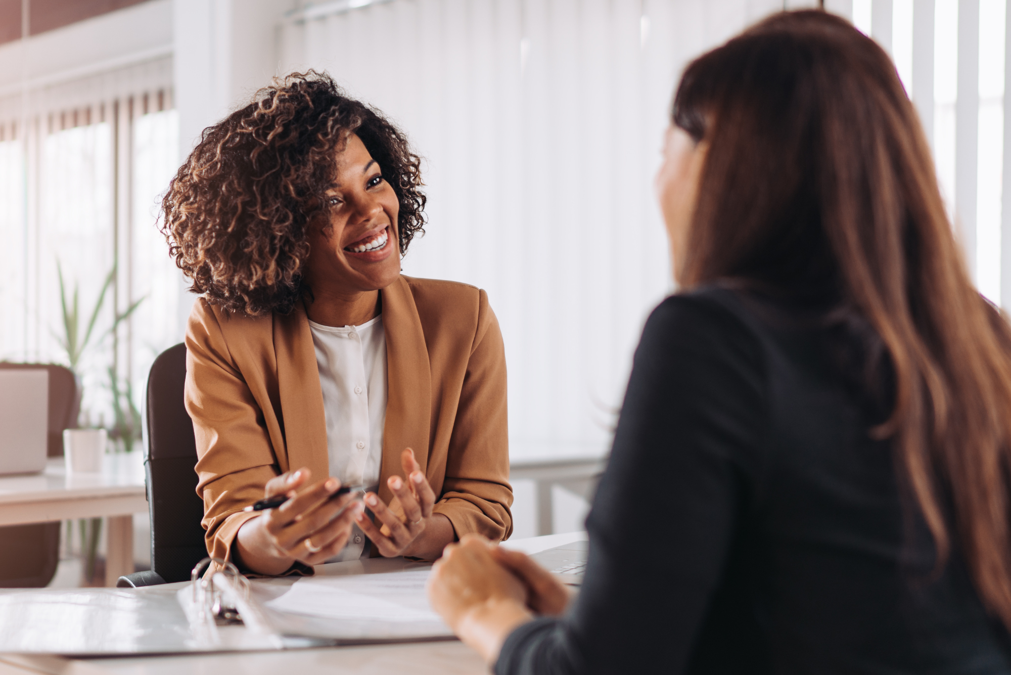 A smiling woman in a tan blazer is sitting at a desk and talking with another woman, illustrating a client vs. customer conversation.