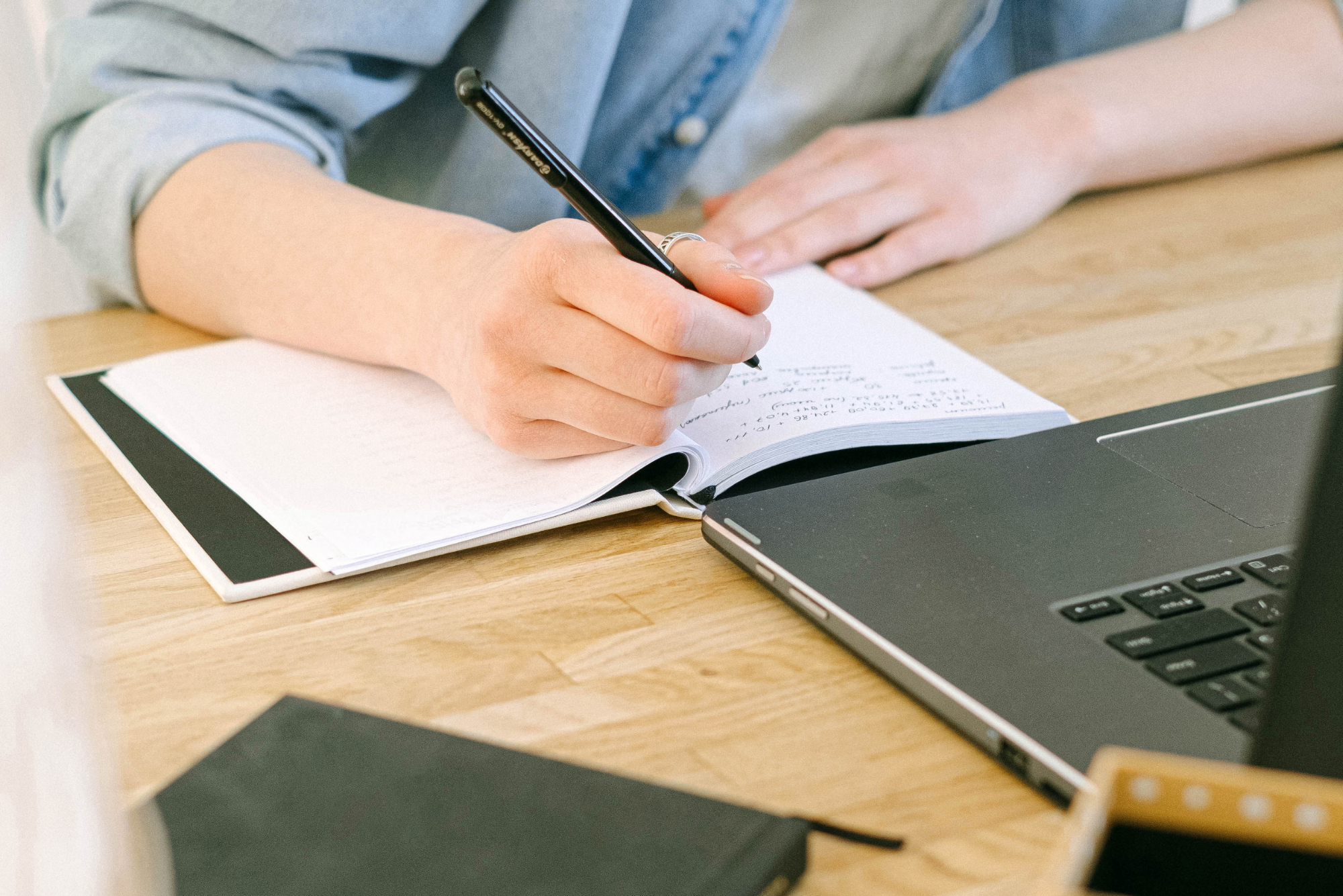 A person writing notes in a notebook at a desk with laptop nearby.