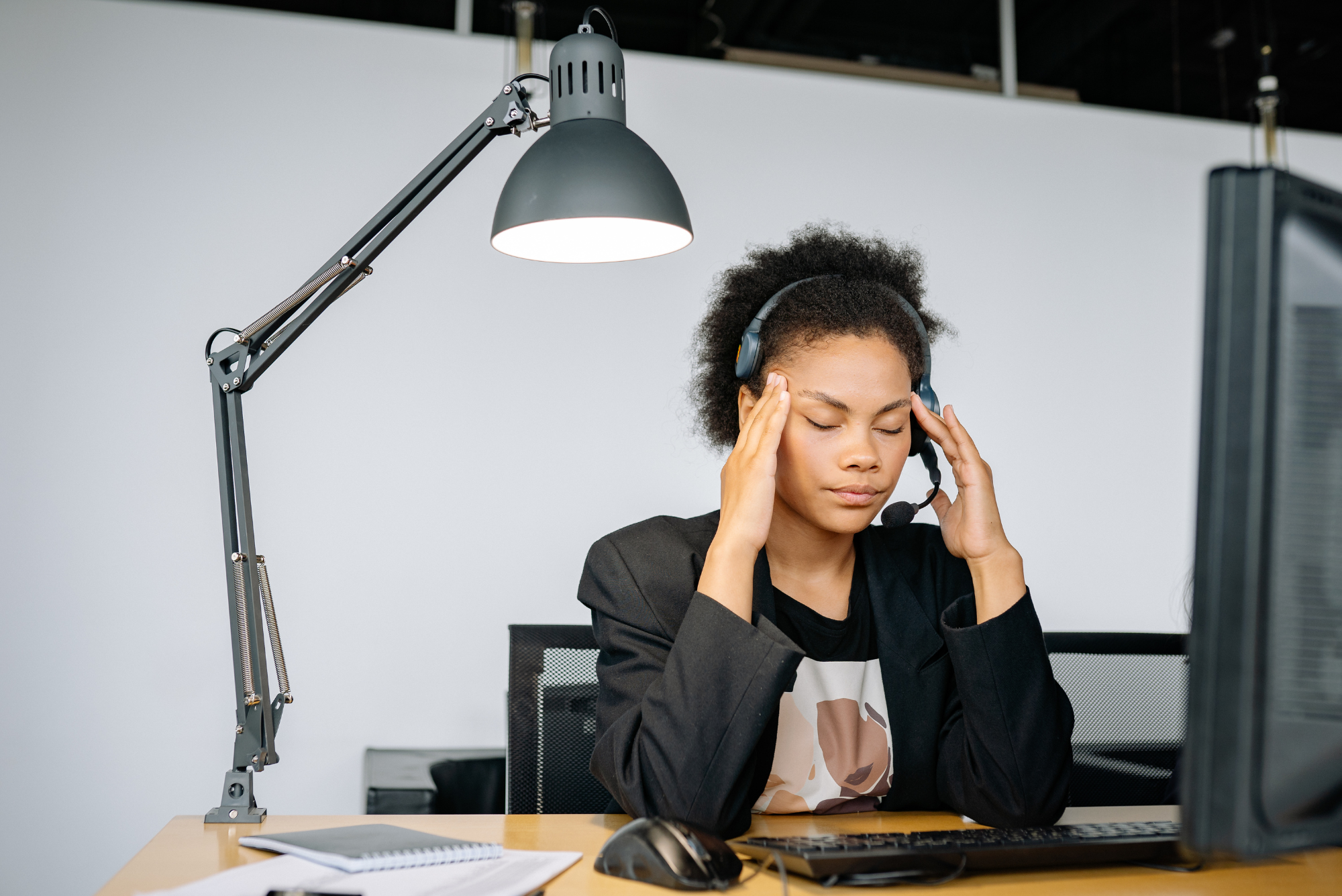 Frustrated employee at a desk rubbing her temples with eyes closed while wearing a headset.