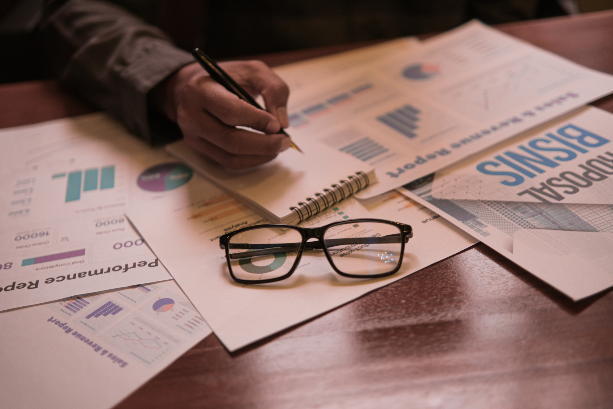 A person writes notes over a desk covered in business reports, charts, and a document labeled “Proposal” with glasses in the foreground.
