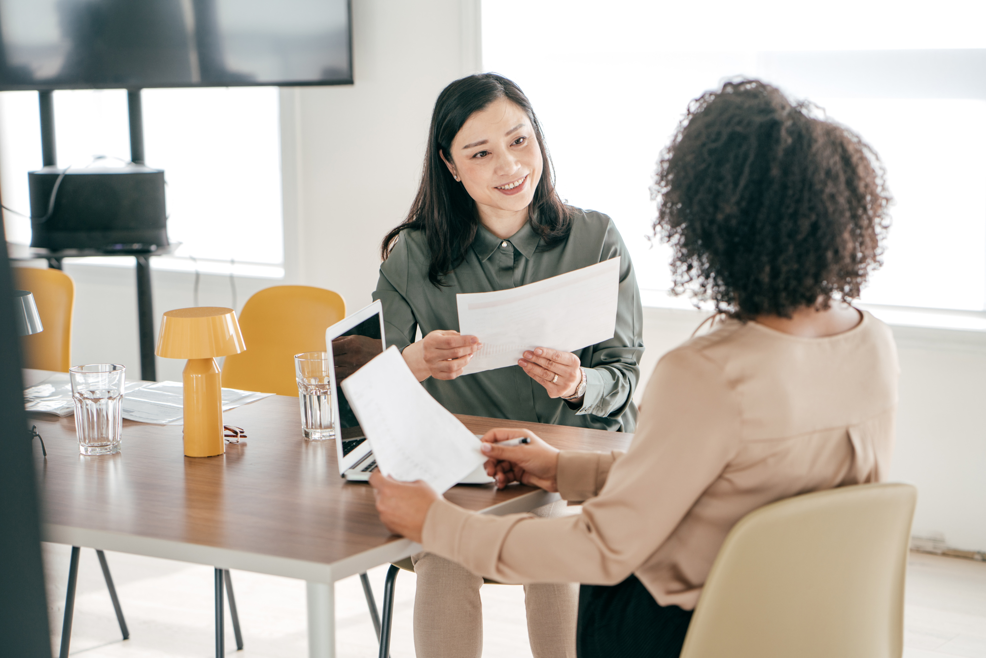 Two women review documents across a table during a job interview in a bright modern office.