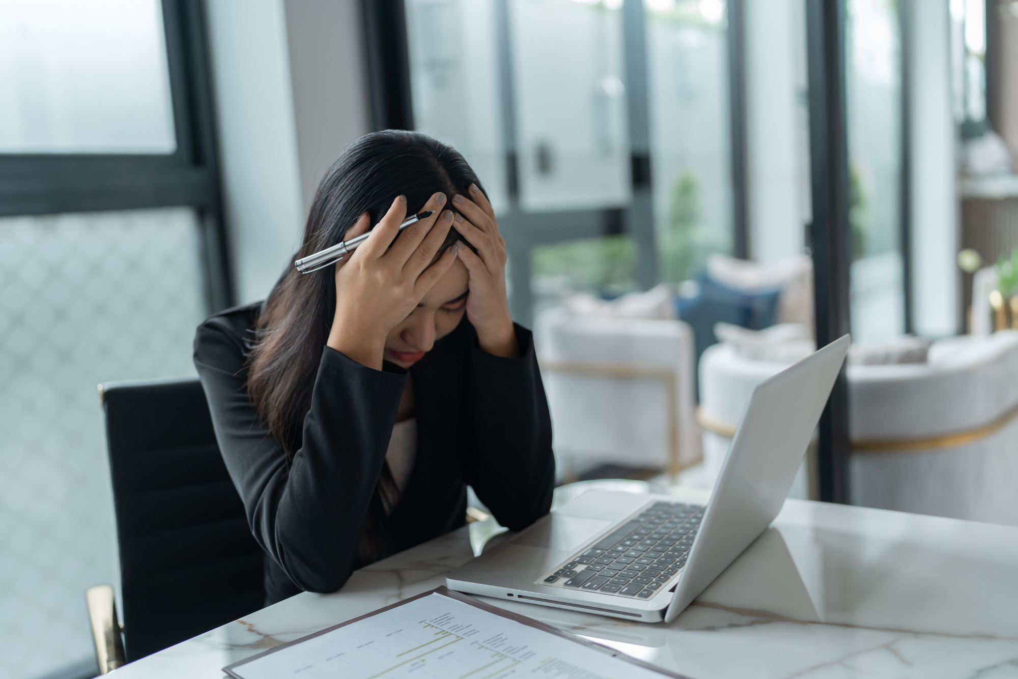 A frustrated businesswoman holds her head in both hands while sitting at a laptop in a bright modern office.