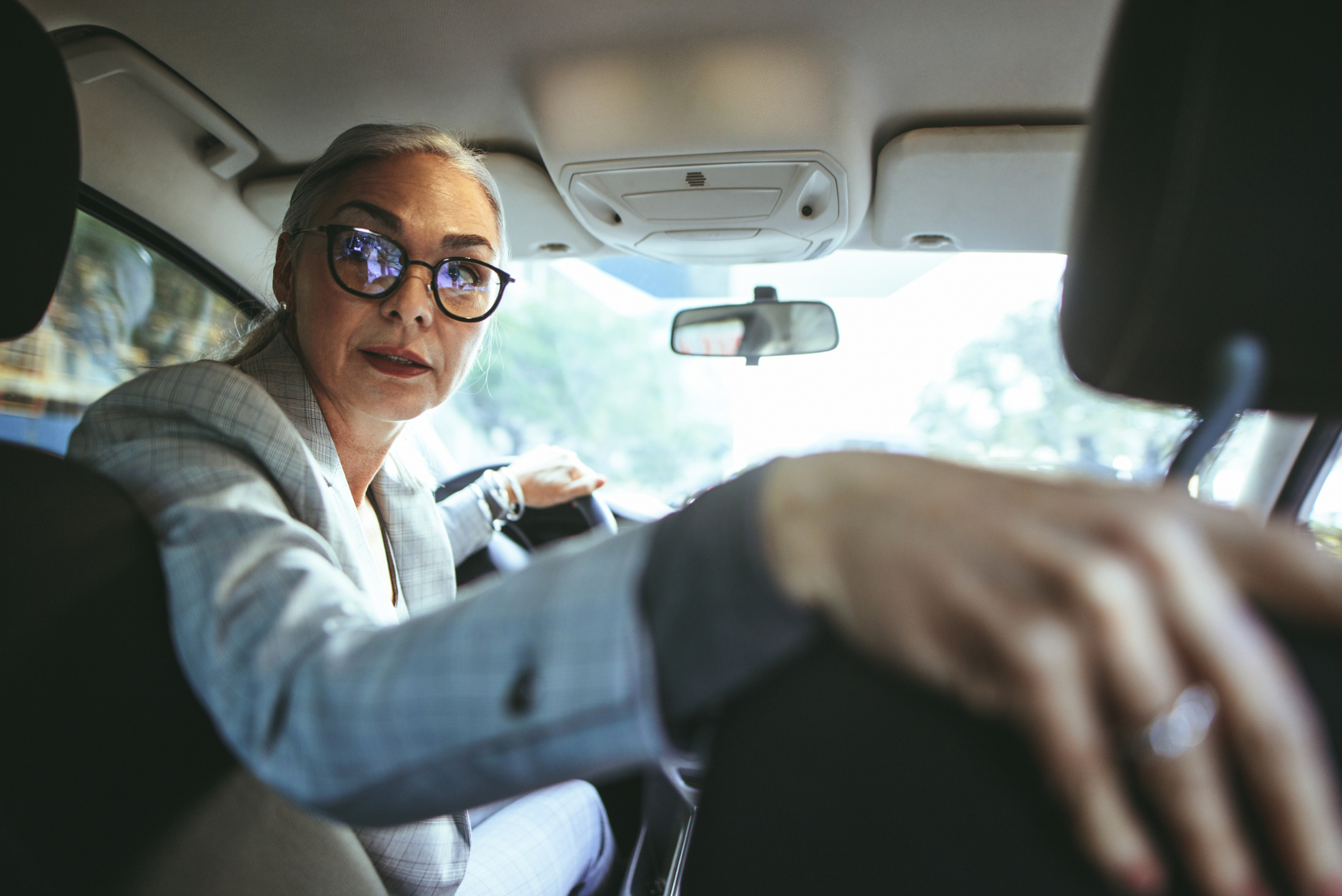 A businesswoman in glasses looks over her shoulder while reversing a car, one arm resting on the passenger seat.