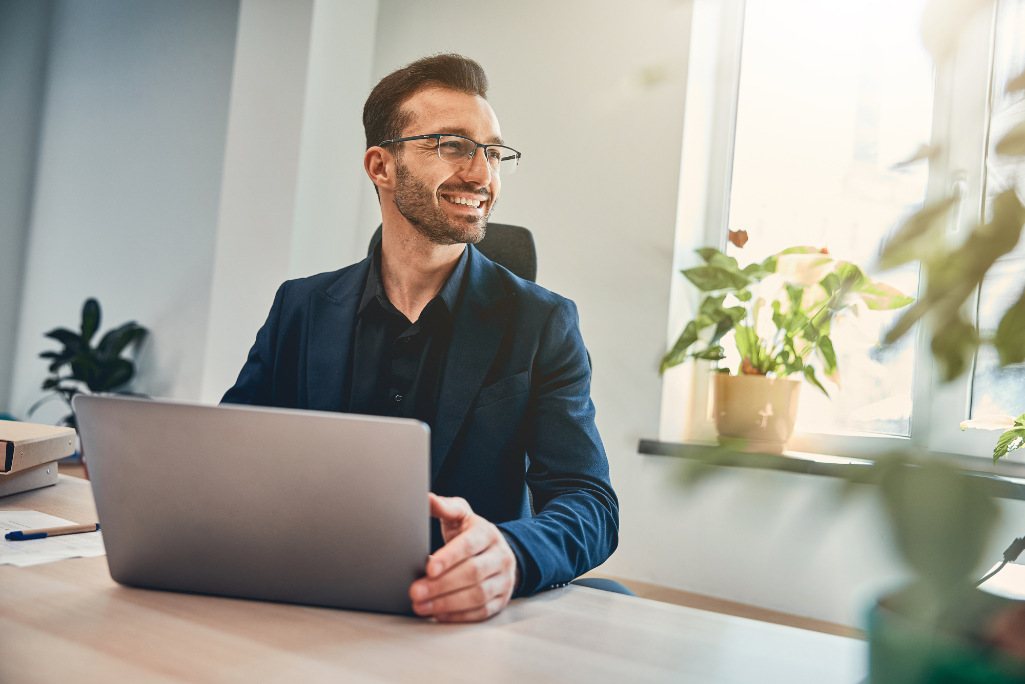 A smiling man in glasses and a navy blazer sits at a laptop in a bright office, looking toward a sunlit window.